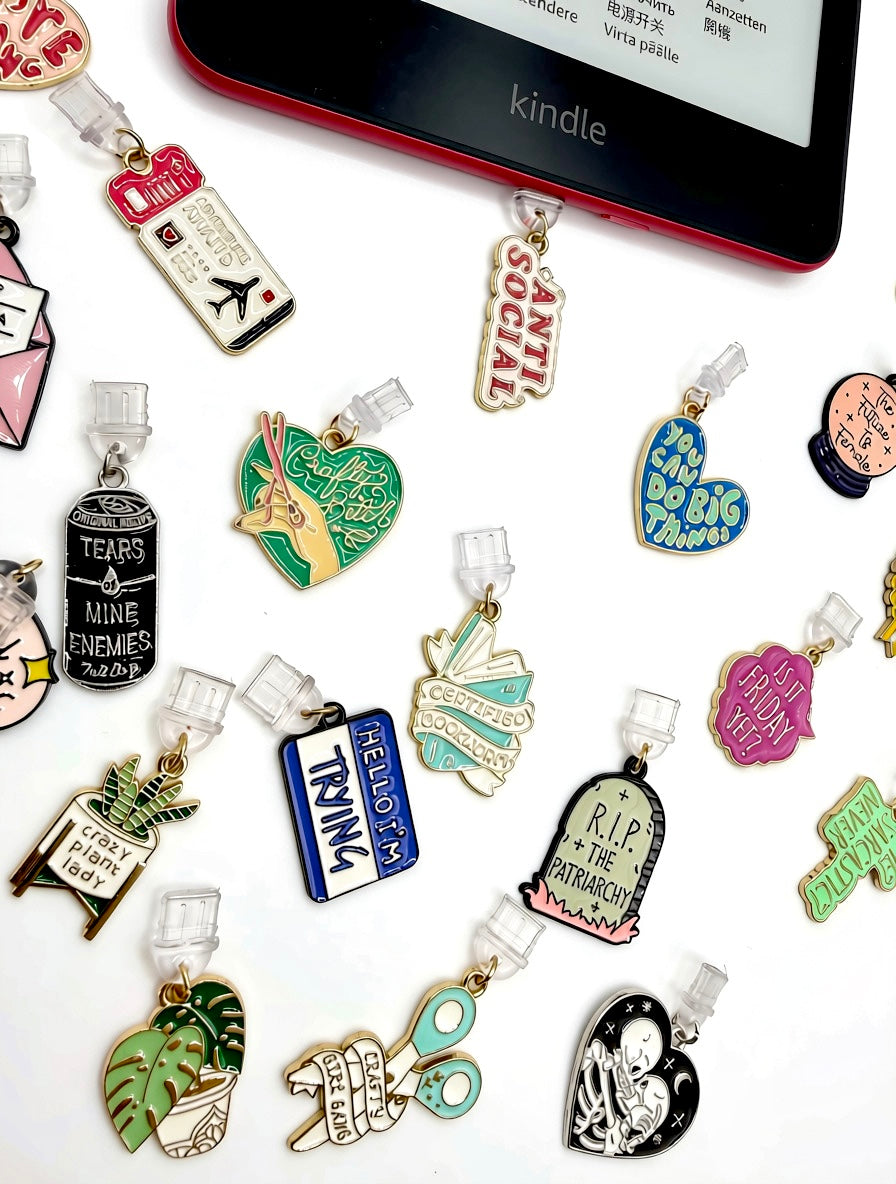 A collection of colorful enamel USB-C Dust Plug Cuver Charms, arranged on a white surface next to a Kindle e-reader. The charms feature various designs including a tombstone reading 'RIP TO THE PATRIARCHY,’ a planter with a snake plant, a book, tropical leaves, and other novelty text-based motifs.